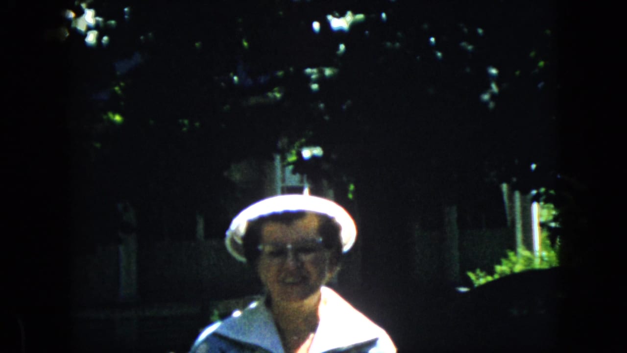 Older woman in a white hat faces the camera with trees and a house behind her.