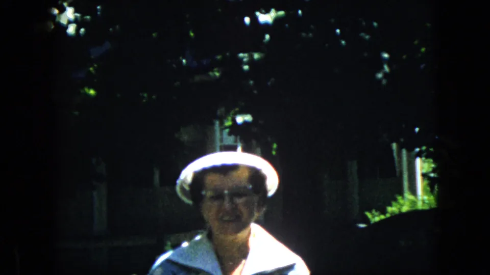 The older woman remains centered in the 1969 California backyard scene, with her white hat catching strong sunlight against deep tree shade and a house facade behind her.