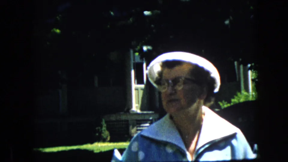 Closer 8mm frame of the older woman in a white hat, glasses, and blue patterned light jacket, with sunlit grass, trees, and a residential exterior visible behind her in California.