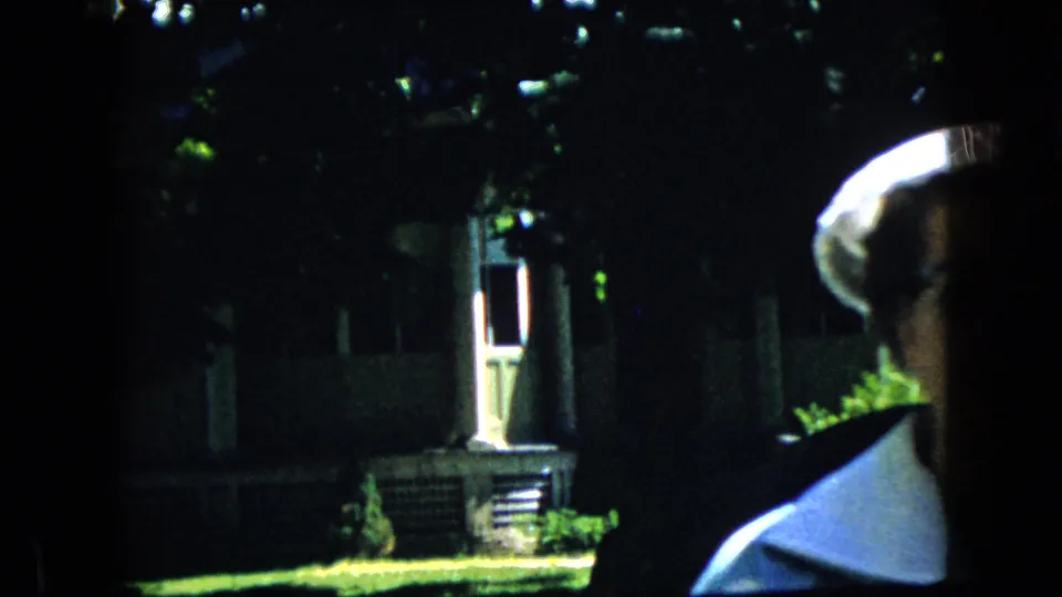 At the end of the clip, the older woman in a white hat and light collared jacket passes close to the camera frame, partly cropped, with a shaded house and garden behind her.