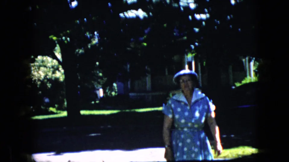 In this California 1969 home-movie frame, an older woman in a blue polka-dot dress and white hat walks toward the camera on a leafy street with lawns and trees behind her.