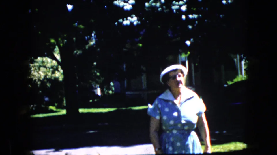 In this California 1969 8mm frame, the older woman is closer to the camera, wearing a blue polka-dot dress with a white collar and hat against a shaded residential backdrop.