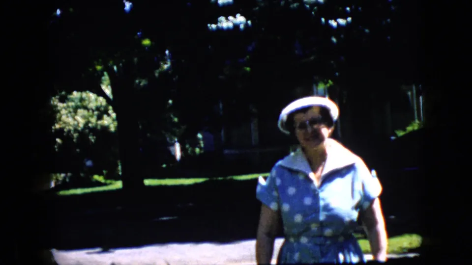 In this California 1969 home-movie frame, the older woman appears nearer and more centered, with dappled sunlight on her blue polka-dot dress and white hat as trees and lawns recede behind her.