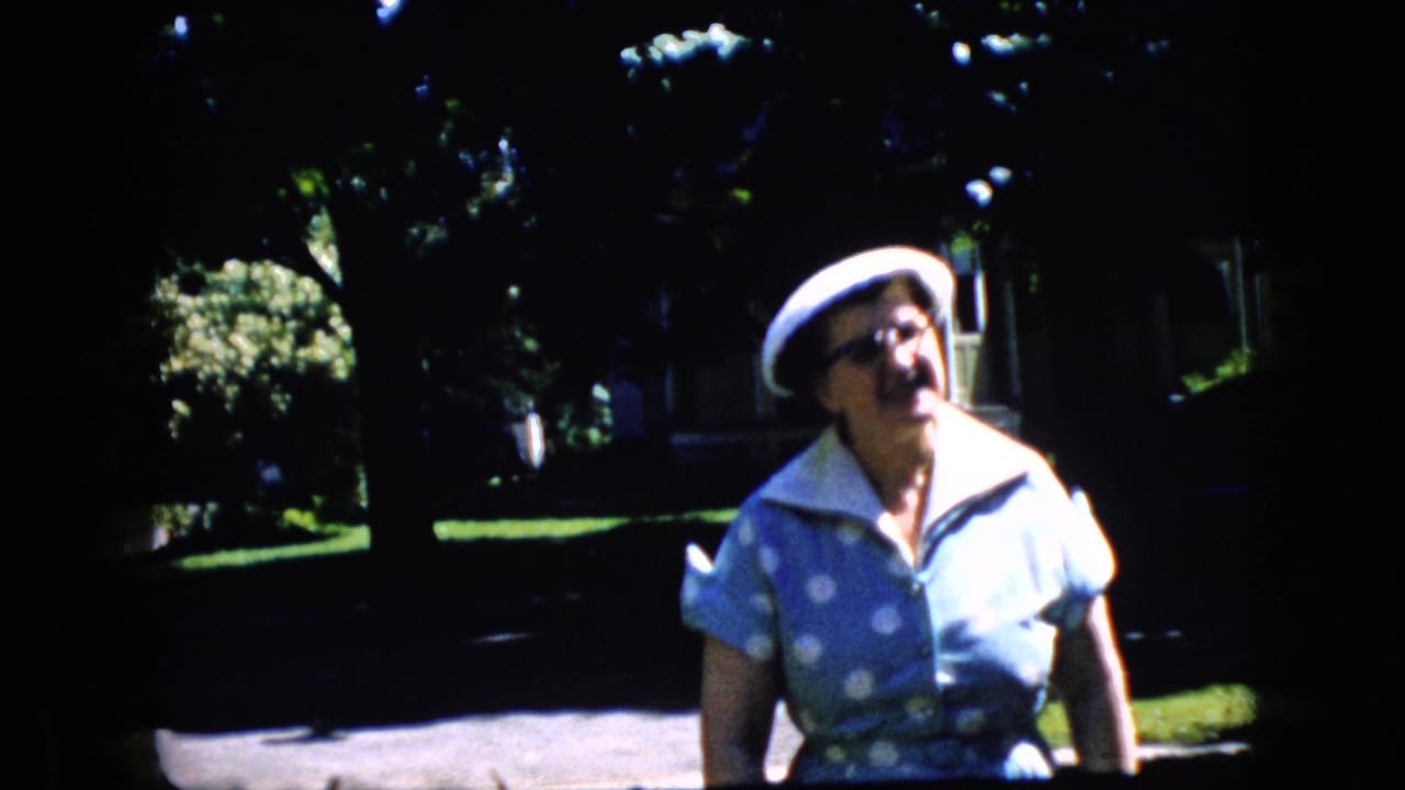 In a 1969 California home-movie frame, an older woman in a blue polka-dot dress, white hat, and glasses walks toward the camera on a sunlit suburban street.