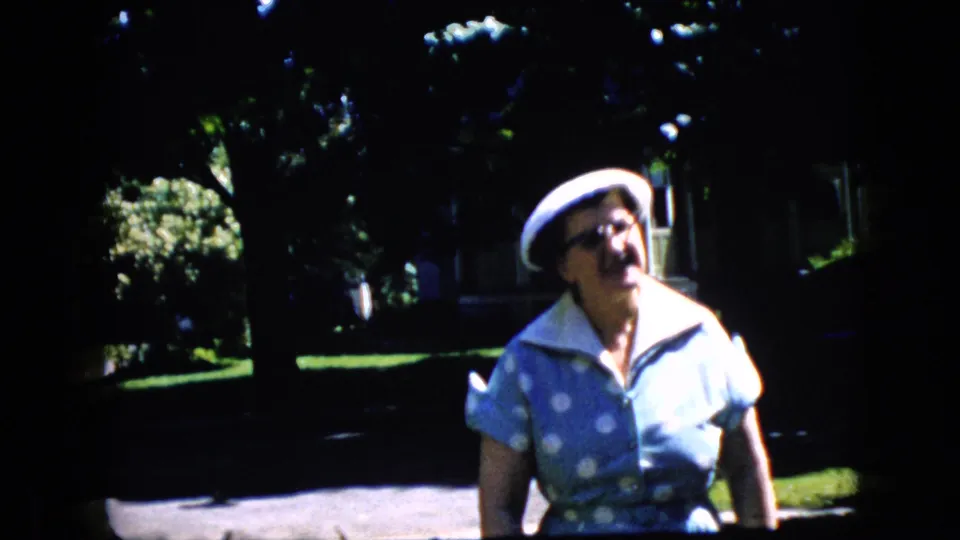 In a 1969 California home-movie frame, an older woman in a blue polka-dot dress, white hat, and glasses walks toward the camera on a sunlit suburban street.