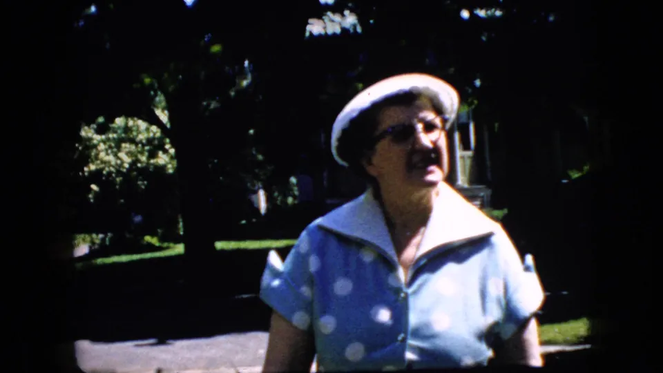 The older woman appears closer to the camera in the 1969 California 8mm footage, wearing glasses, a white hat, and a blue polka-dot dress against a shaded residential backdrop.