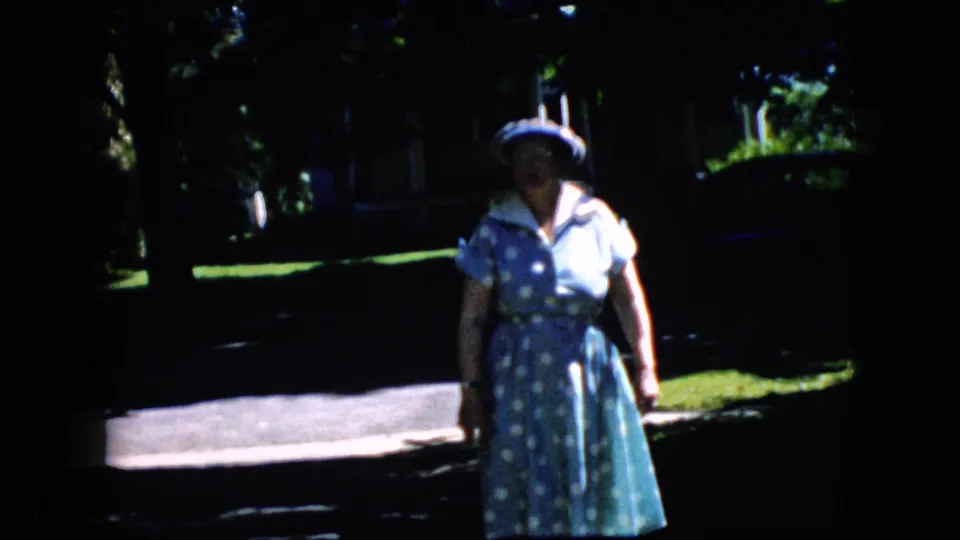 A wider 1969 California home-movie frame shows the older woman in her blue polka-dot dress and white hat standing or moving slowly on the residential street with lawns and trees behind her.