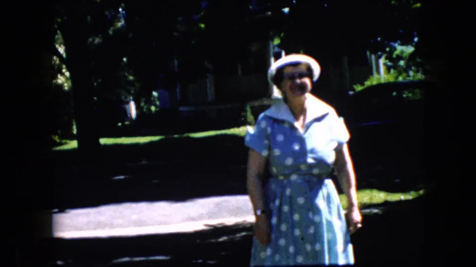 At the end of the 1969 California clip, the older woman faces the camera in a blue polka-dot dress and white hat, with bright pavement, lawns, and shaded houses behind her.