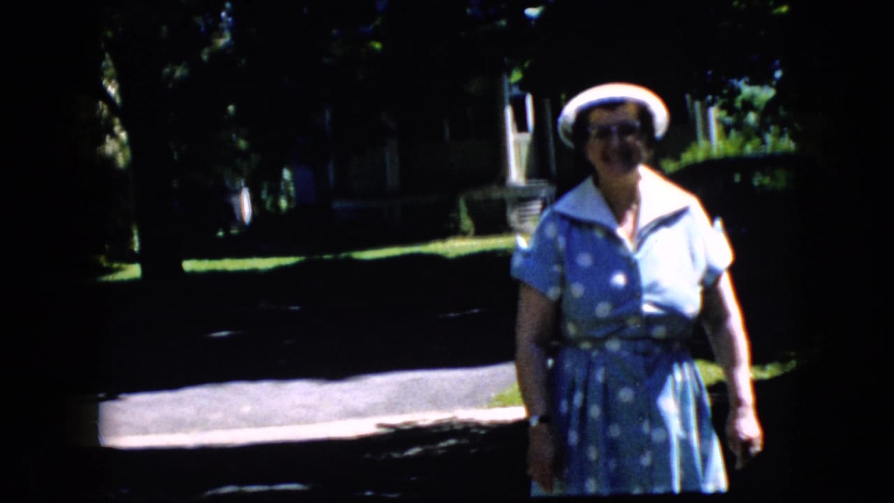 Older woman in a blue polka-dot dress and white hat standing on a shaded sunny street.