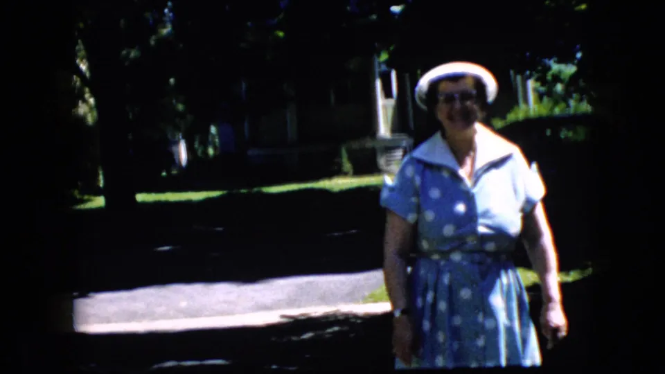 In a 1969 California home-movie frame, an older woman in glasses, a white hat, and a blue polka-dot dress faces the camera on a shaded suburban street.
