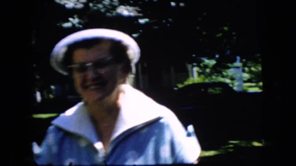 A close handheld frame shows the older woman smiling as she approaches, with her white hat, glasses, pale collar, and blue dress prominent.