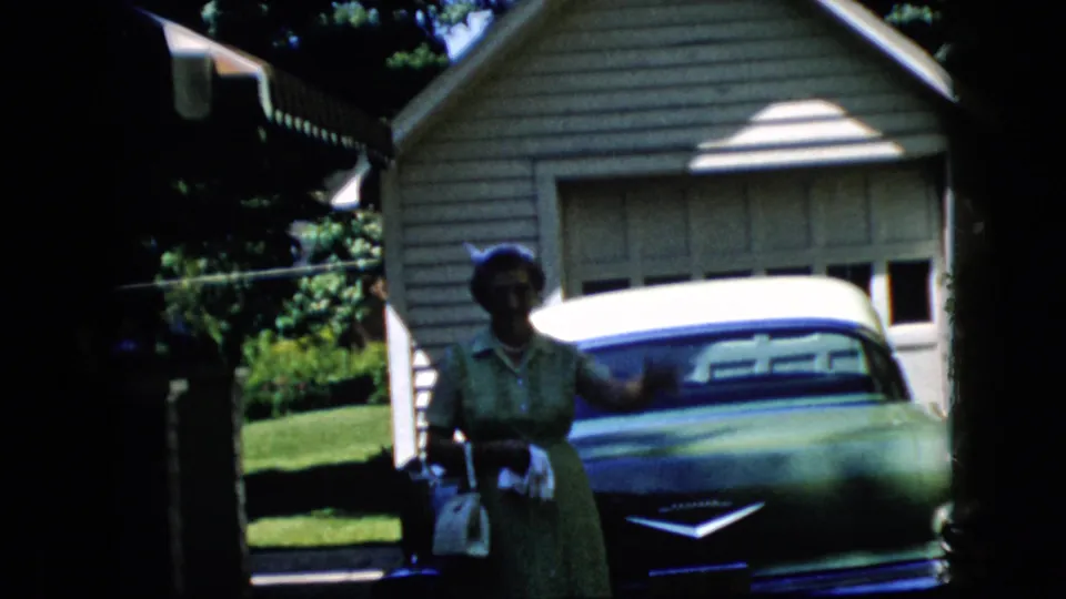 The woman in a green dress begins a small wave while holding a handbag beside a classic car in a sunny suburban driveway.
