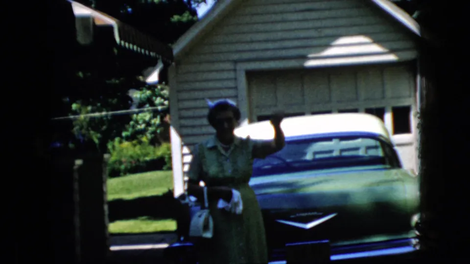 The woman waves toward the camera while standing beside the parked classic car, framed by a garage, house exterior, lawn, and deep shadow at the edge of the film frame.