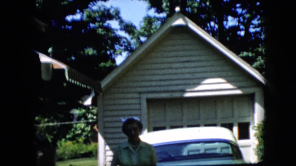 The woman remains near the green classic car and detached garage, with the 8mm frame cropping her lower in the image.