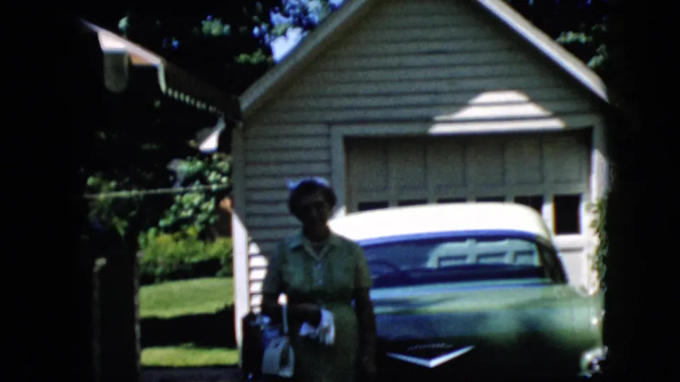 The elderly woman faces the camera with a white handbag at her side, standing before a green car and suburban garage in the California home-movie scene.