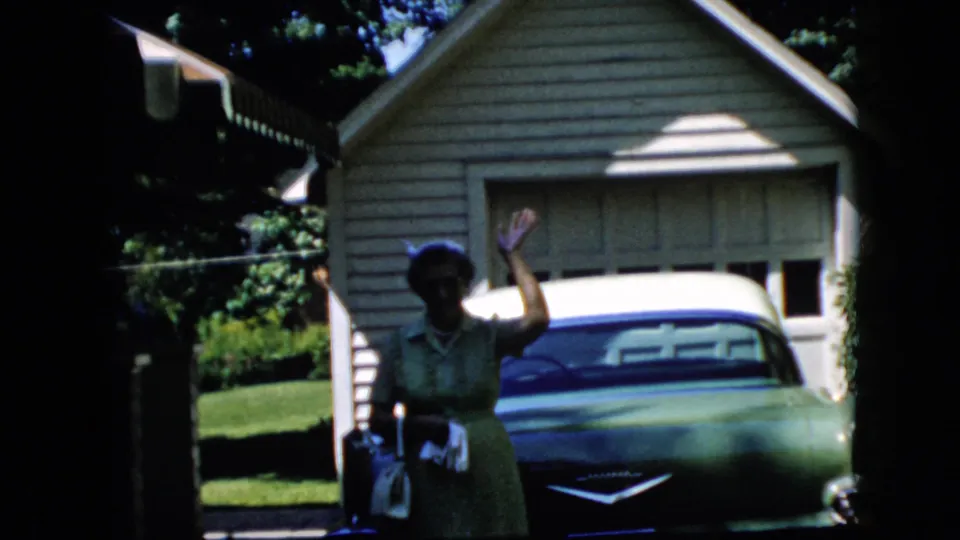 The woman raises one hand in a wave toward the camera while standing beside the green classic car outside the suburban garage.