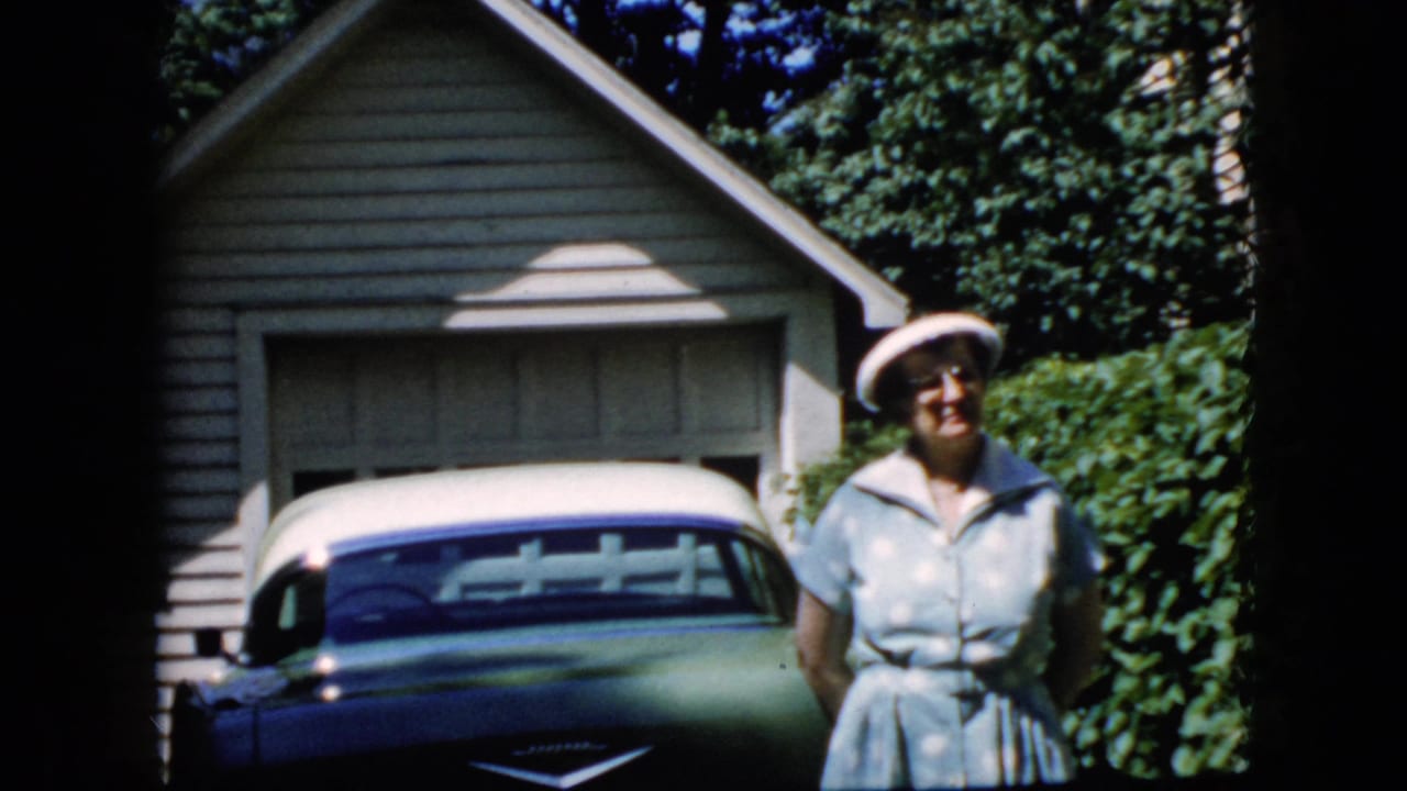 Smiling woman in a light dress and white hat standing beside a classic car near a garage.