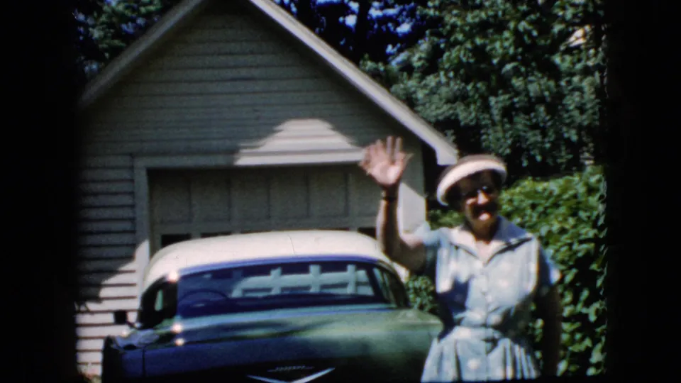 A woman in a light blue dress and white hat waves toward the camera while standing beside a classic car in a sunny California residential driveway setting.