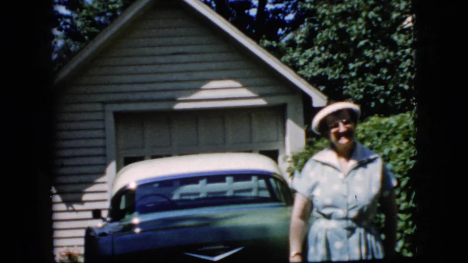 A woman smiles beside a parked green-and-white classic car, with a detached garage, hedges, and trees visible in the 1969 California home-movie frame.