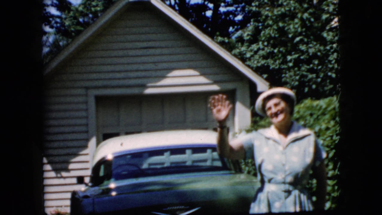 Older woman waves beside a blue and white classic car in front of a garage.