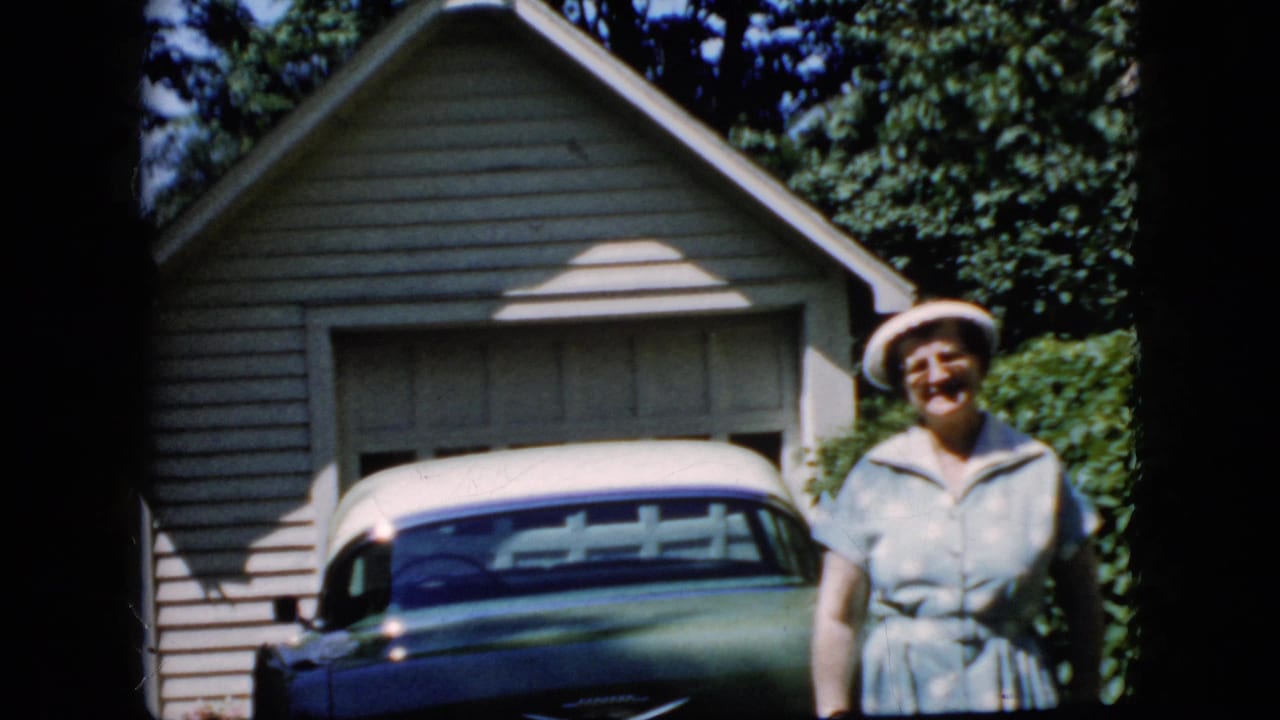 Older woman smiles beside a classic car parked in front of a garage.