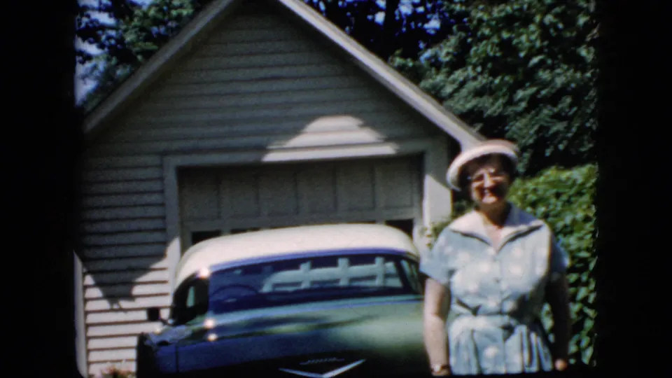 The woman stands facing the camera beside the parked blue and white car, framed by a beige-sided garage, green hedge, and heavy 8mm frame-edge shadowing.