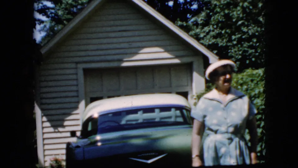 The woman turns slightly to the side while standing next to the blue and white car in the suburban driveway, with the garage, hedge, and trees still visible.