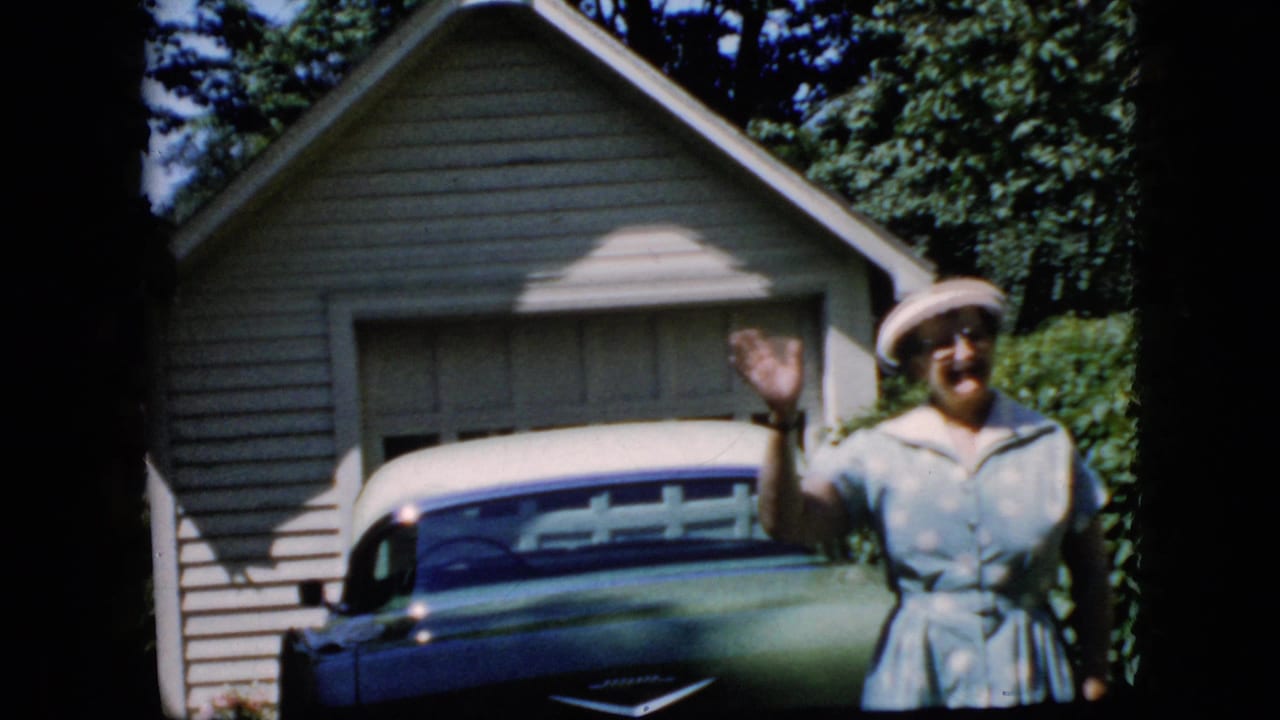 Elderly woman waves beside a green and white car in front of a garage.