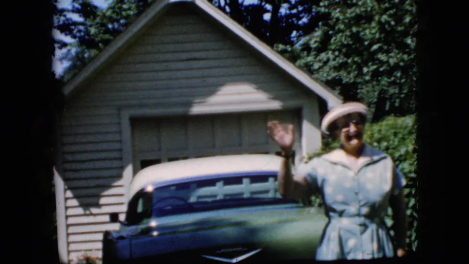 In a 1969 California home-movie frame, an elderly woman in a light blue dress and white hat waves beside a green and white car parked at a suburban garage.