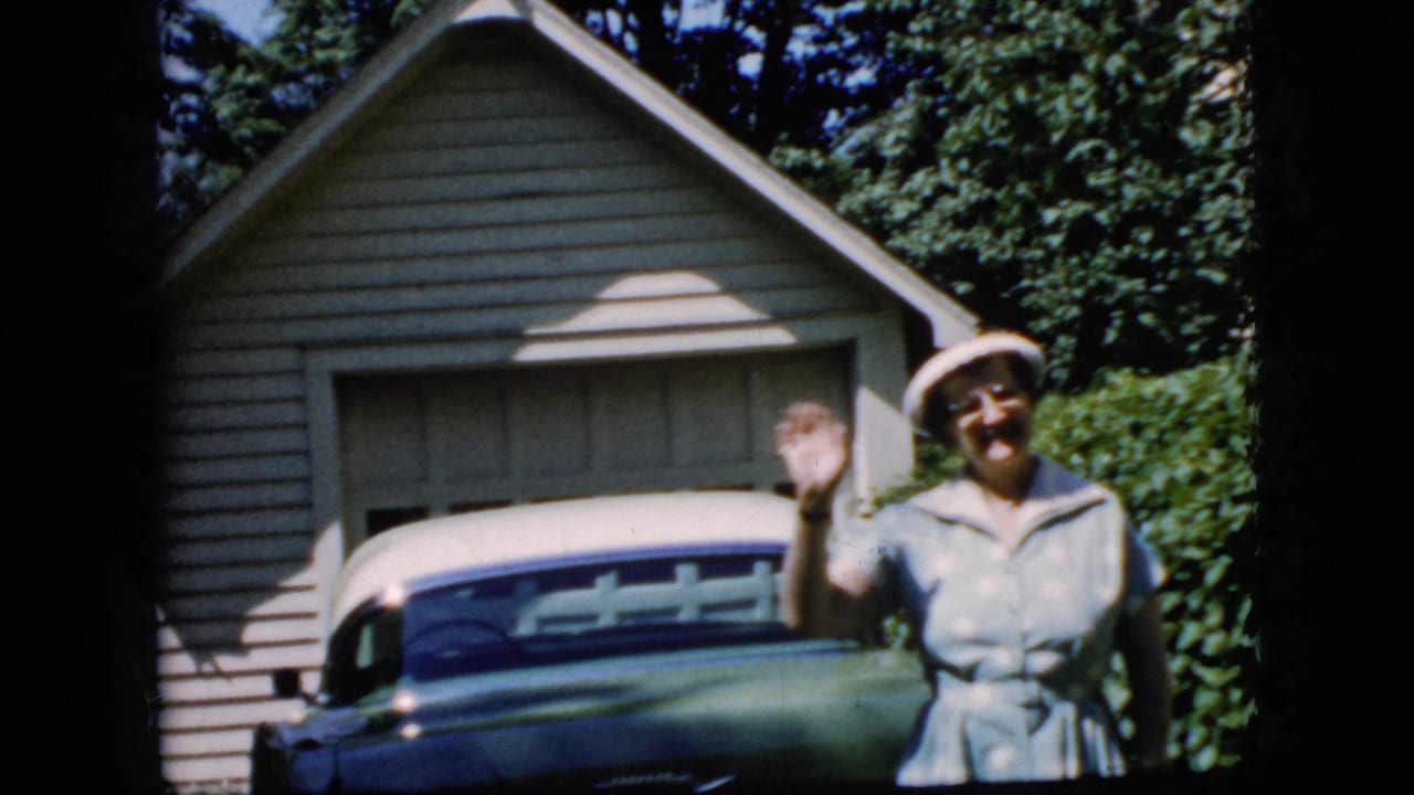Woman continues waving near a parked car and garage in bright sunlight.