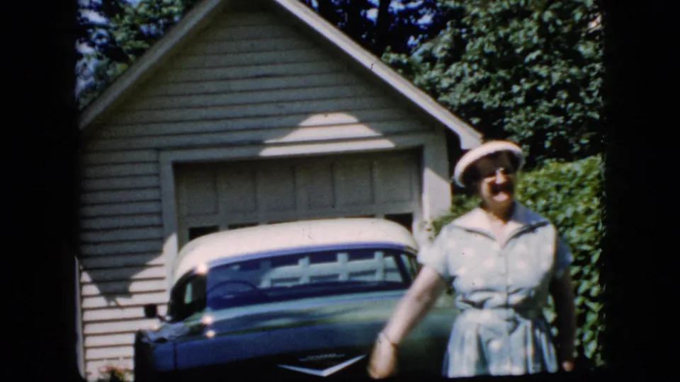 The woman smiles with her arm lowered after waving, standing beside the classic car in a sunny suburban driveway setting.
