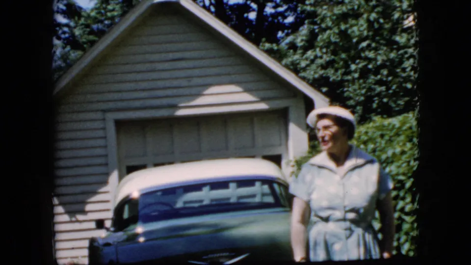 At the end of the clip, the woman turns slightly to the side beside the green and white car, with the garage and leafy shrubs behind her.