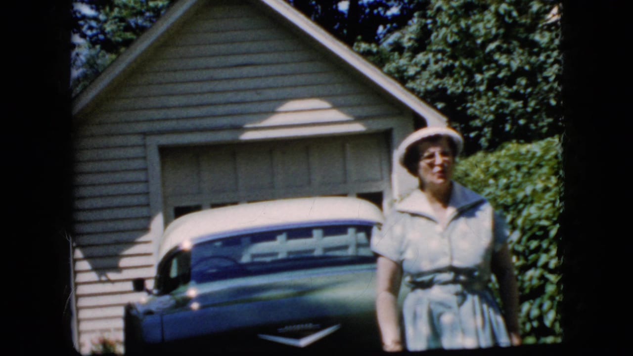 Elderly woman in a light dress and white hat standing beside a blue and white car outside a garage.
