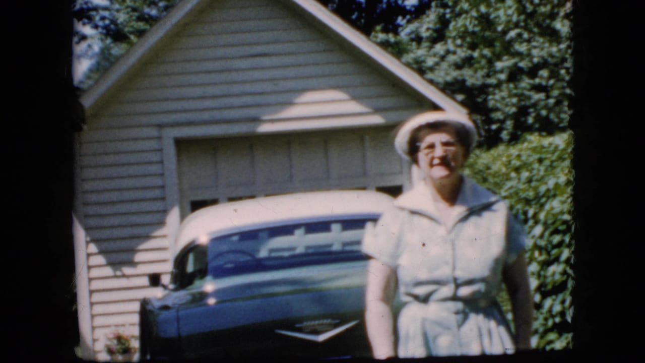 Smiling elderly woman standing closer to camera with a parked car and garage behind her.