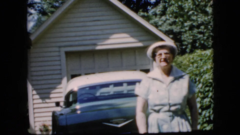 The woman stands closer in the frame, smiling in a white hat and light dress with the blue and white car partly visible behind her.