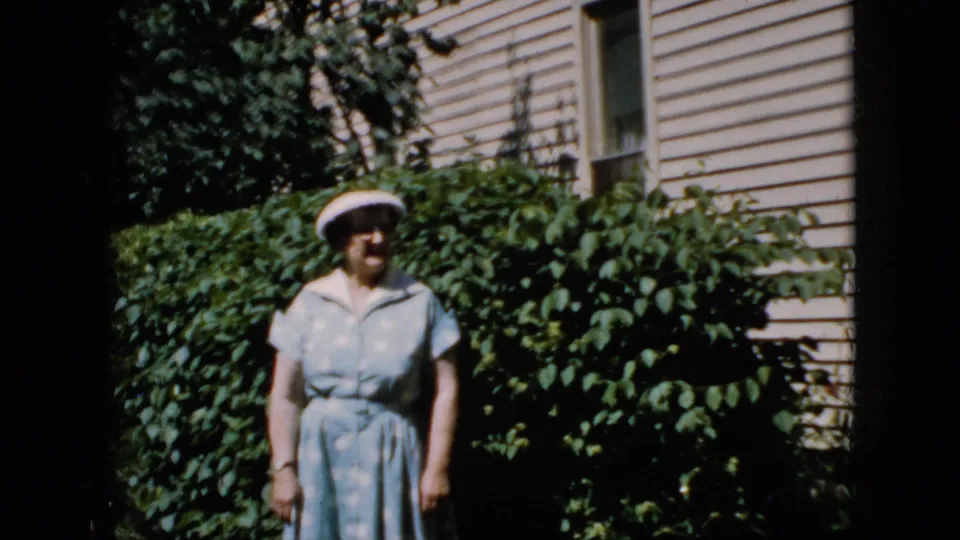 The woman appears lower in the frame beside dense green hedges, with beige house siding and a window visible behind her.