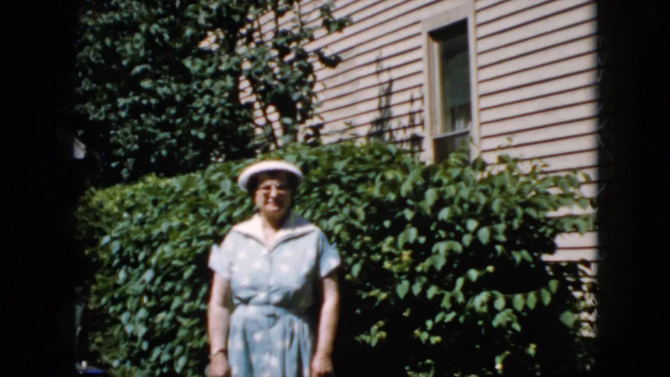 The elderly woman faces the camera in front of tall hedges and a beige-sided house, with strong sunlight and shadow across the frame.