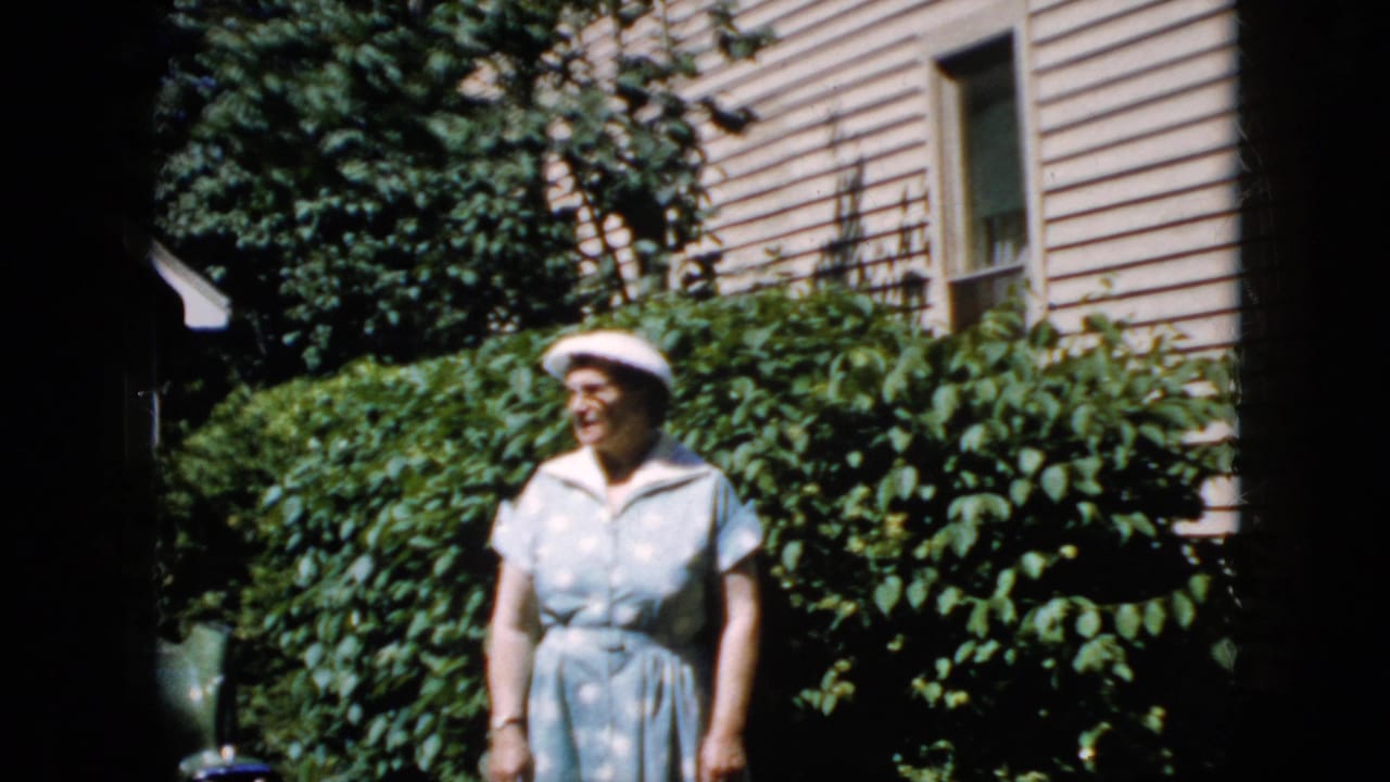 In the 1969 California home-movie clip, an elderly woman in a pale blue dress and white hat stands near dense greenery outside a beige-sided house.