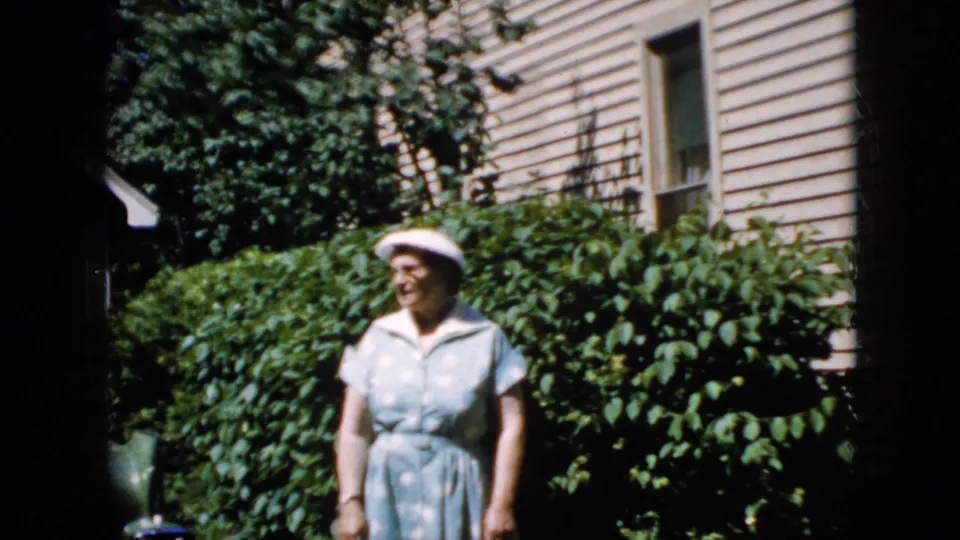 In the 1969 California home-movie clip, an elderly woman in a pale blue dress and white hat stands near dense greenery outside a beige-sided house.