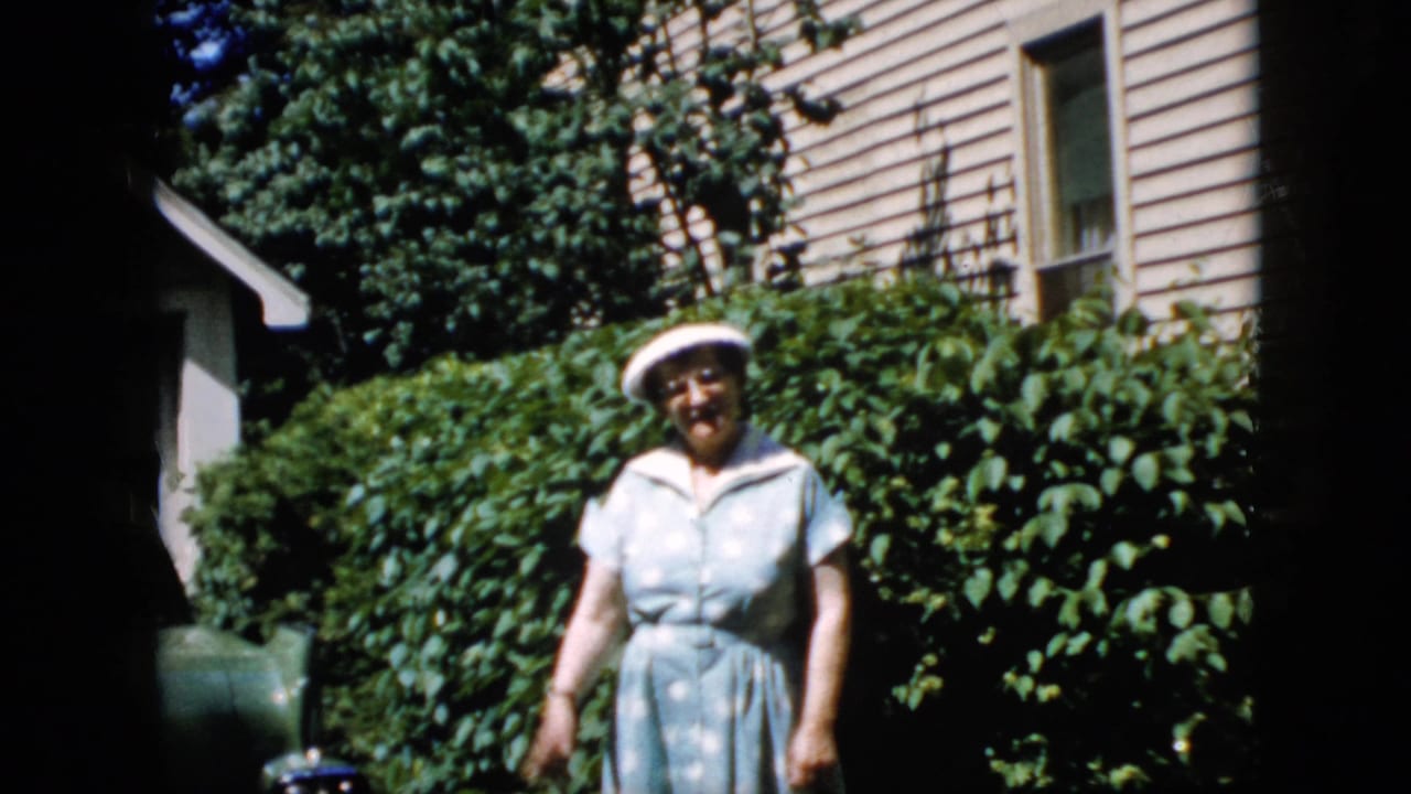 The woman faces the camera in a California residential yard, framed by leafy shrubs, beige siding, and strong sunlight.