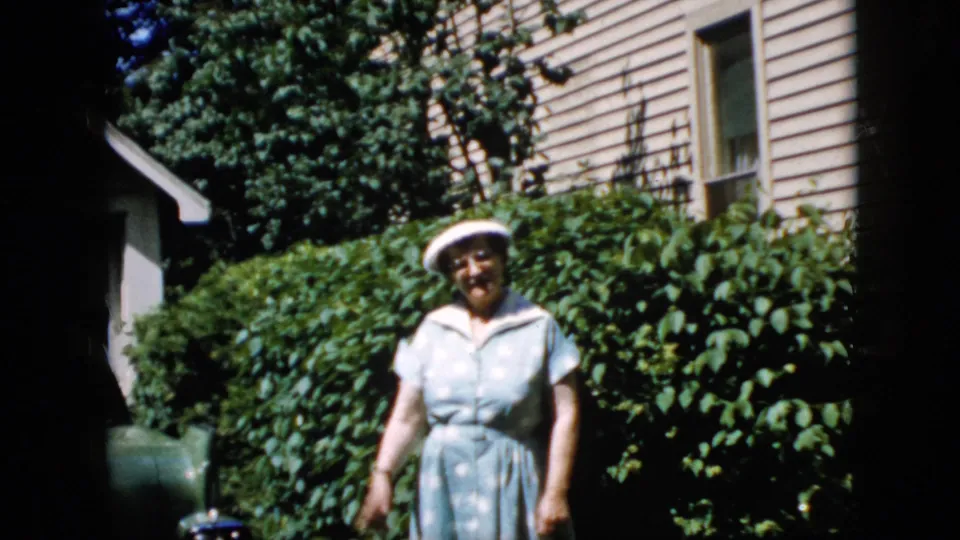 The woman faces the camera in a California residential yard, framed by leafy shrubs, beige siding, and strong sunlight.