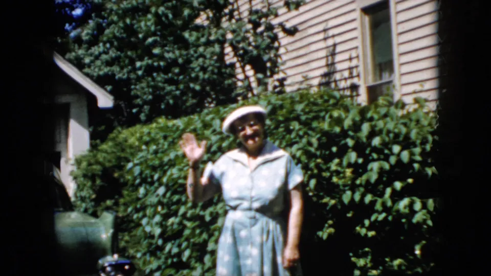 The woman smiles and waves toward the camera, standing in front of shrubs and a house exterior in the 1969 California home-movie scene.