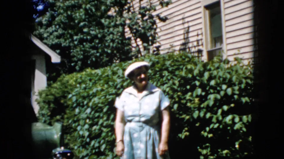 At the end of the clip, the woman stands relaxed in the yard with the beige house, window, and dense shrubs still visible behind her.