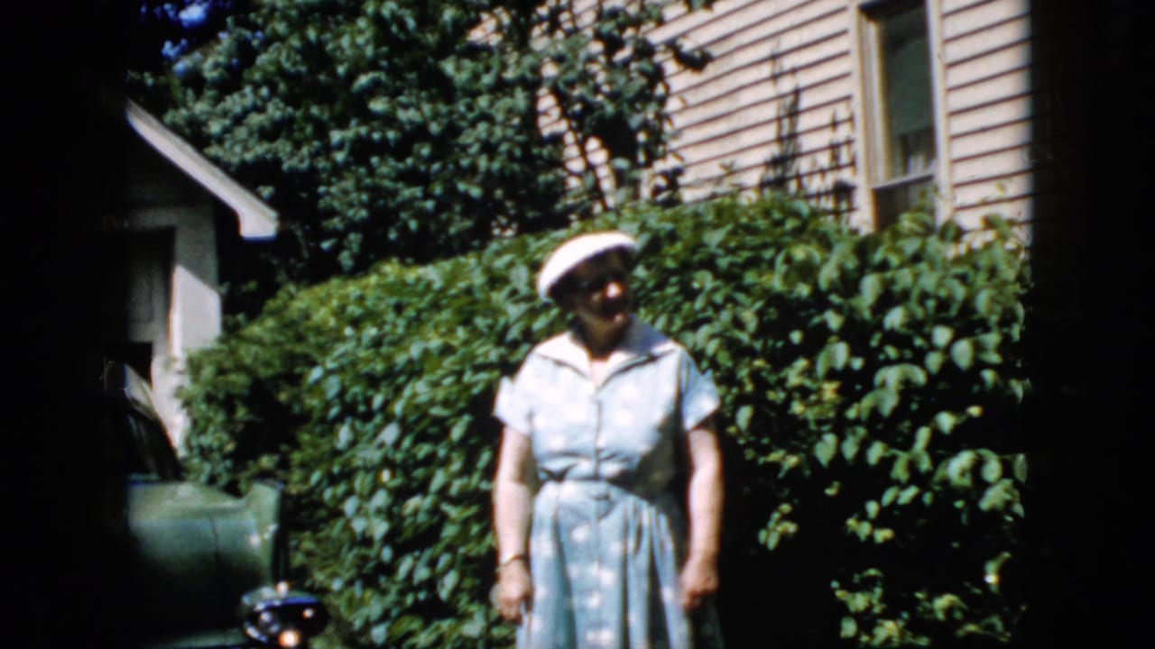 In this 1969 California 8mm home-movie frame, an older woman in a light blue dress and white hat faces the camera in a sunlit backyard with shrubs, house siding, and part of a car visible.