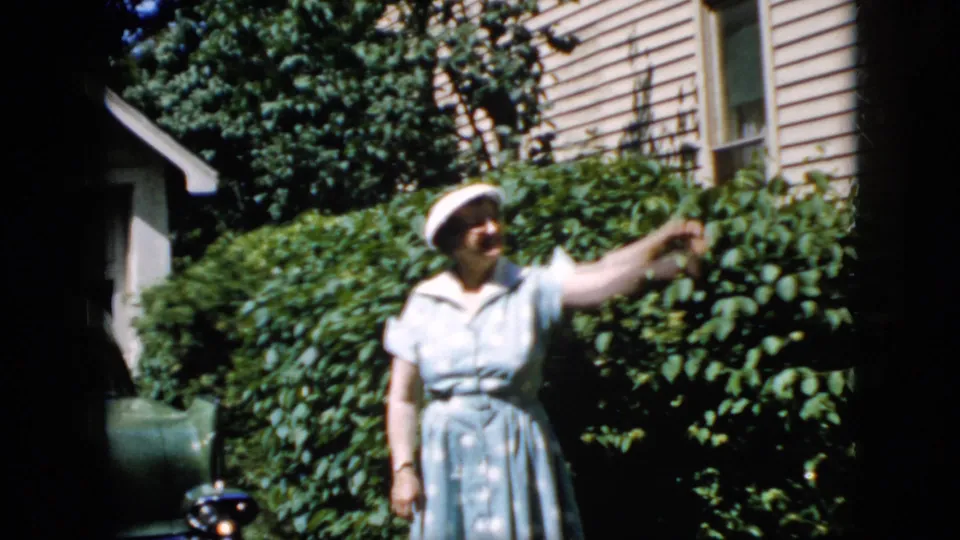 The older woman smiles toward the camera and lifts her arm in a friendly wave in a 1969 California backyard, with green shrubs, house siding, and a partial car along the left edge.