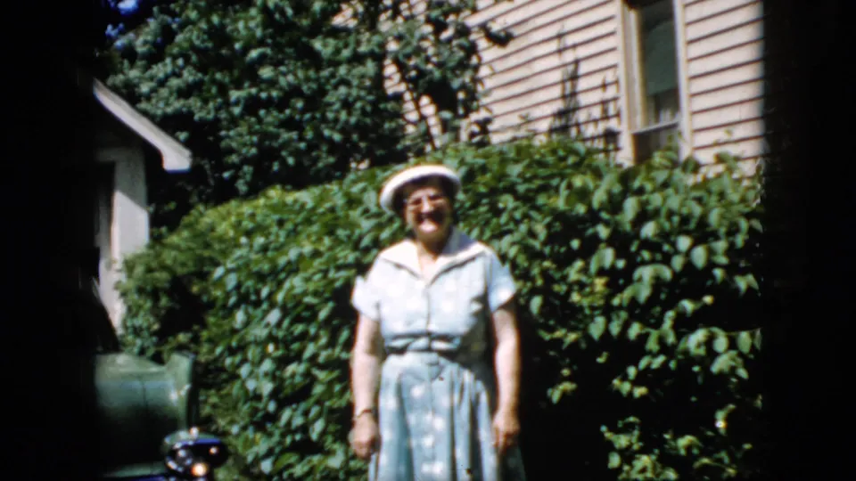 The woman returns her arm downward and stands facing the home-movie camera in the California backyard, framed by leafy shrubs, beige siding, a window, and bright daylight.