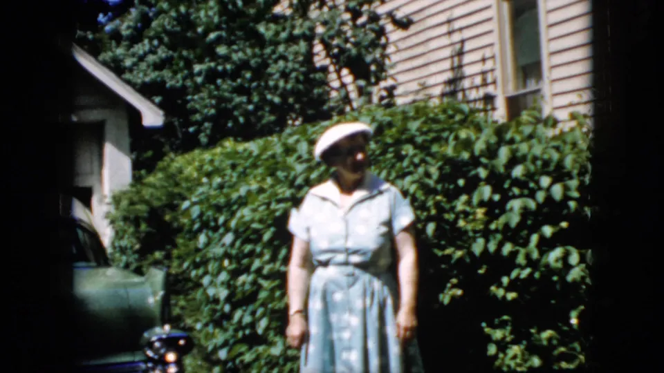 At the end of the clip, the older woman stands in the sunlit backyard and looks slightly off camera, with shrubs, beige house siding, and a dark green car edge visible nearby.