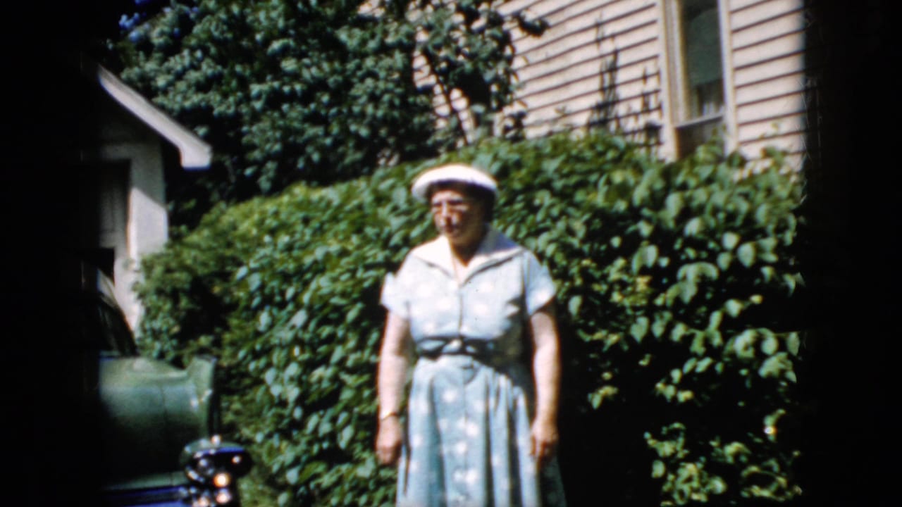 Woman in a light dress and white hat standing near shrubs and a parked car.