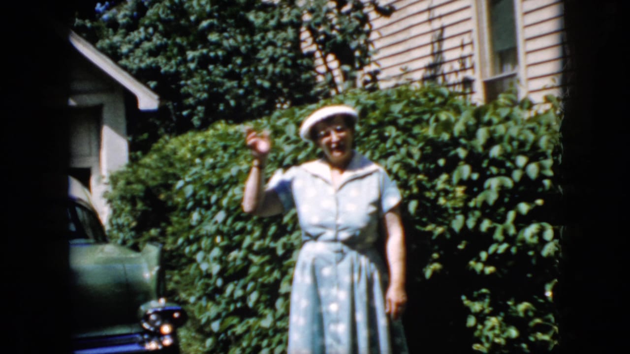 Woman smiling and waving beside tall shrubs in front of a house.
