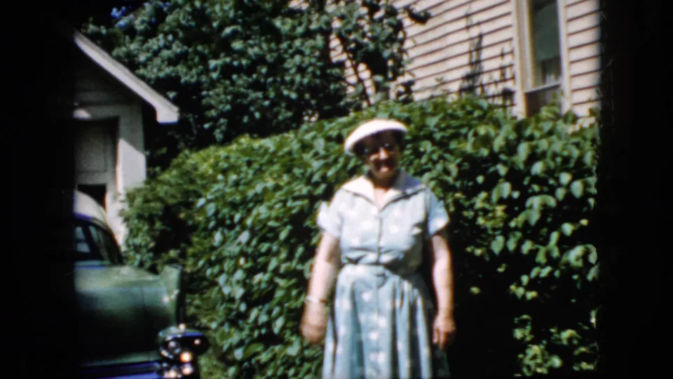 The woman stands closer in the 1969 California home-movie frame, with her white hat, light dress, leafy shrubbery, and part of a parked car visible around her.